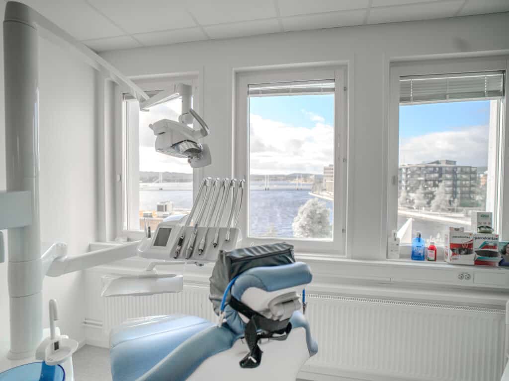 "Behandlingsrum på Smile Tandvård med en tandläkarstol och utrustning, stora fönster som släpper in naturligt ljus och utsikt över staden.