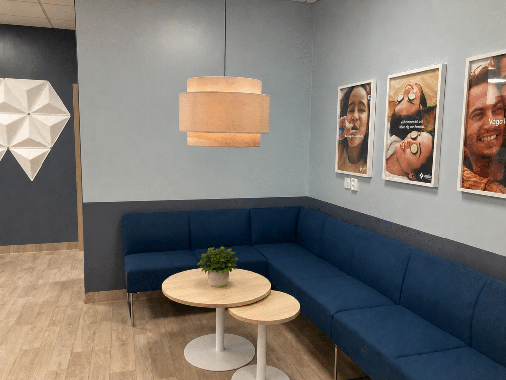Väntrum på Smile Tandvård Karlskrona med blå hörnsoffa, två runda träbord varav det större har en grön växt, samt ljusa väggar med patientbilder.