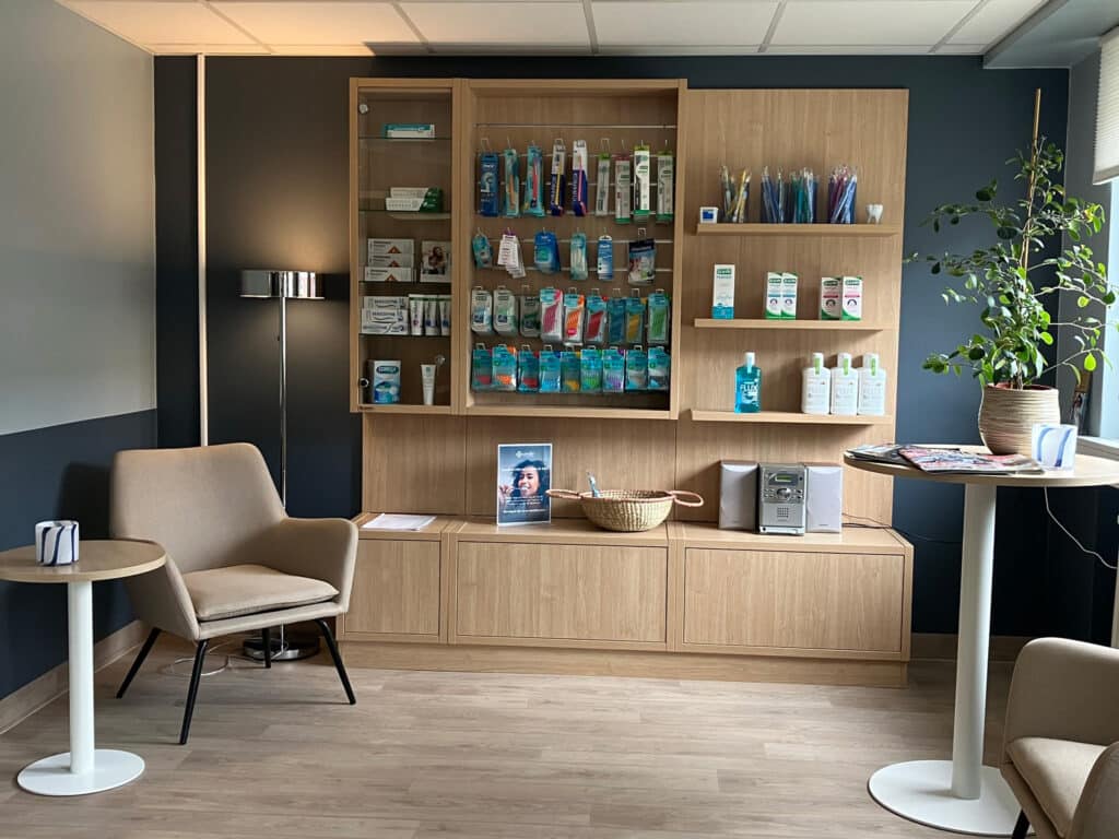 Elegant väntrum hos Smile Tandvård Karlstad, med en stilren träinredning och hyllor fyllda med tandvårdsprodukter. Bekväma fåtöljer och små runda bord skapar en avslappnad atmosfär, medan gröna växter bidrar till en fräsch och välkomnande känsla.