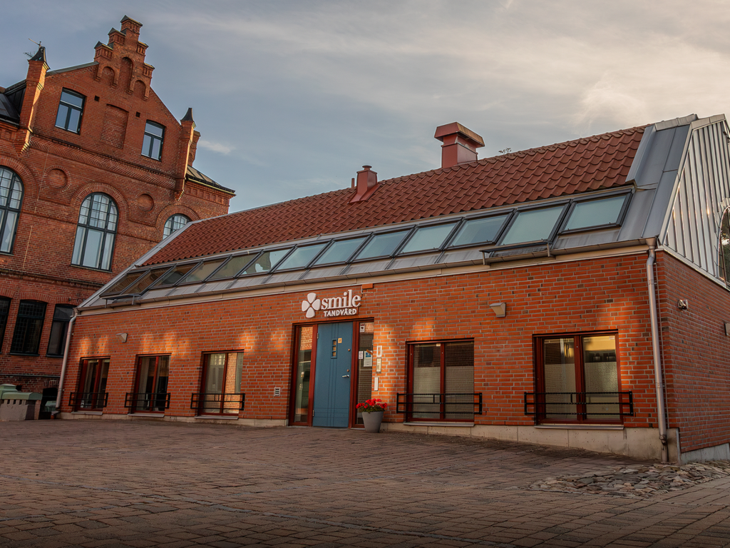 Smile Tandvård i Lund med röd tegelfasad och moderna glaspartier. Byggnaden har en industriell design med en lutande takstruktur, omgiven av äldre arkitektur i bakgrunden. Solen är på väg ner och skapar en mjuk belysning över platsen.