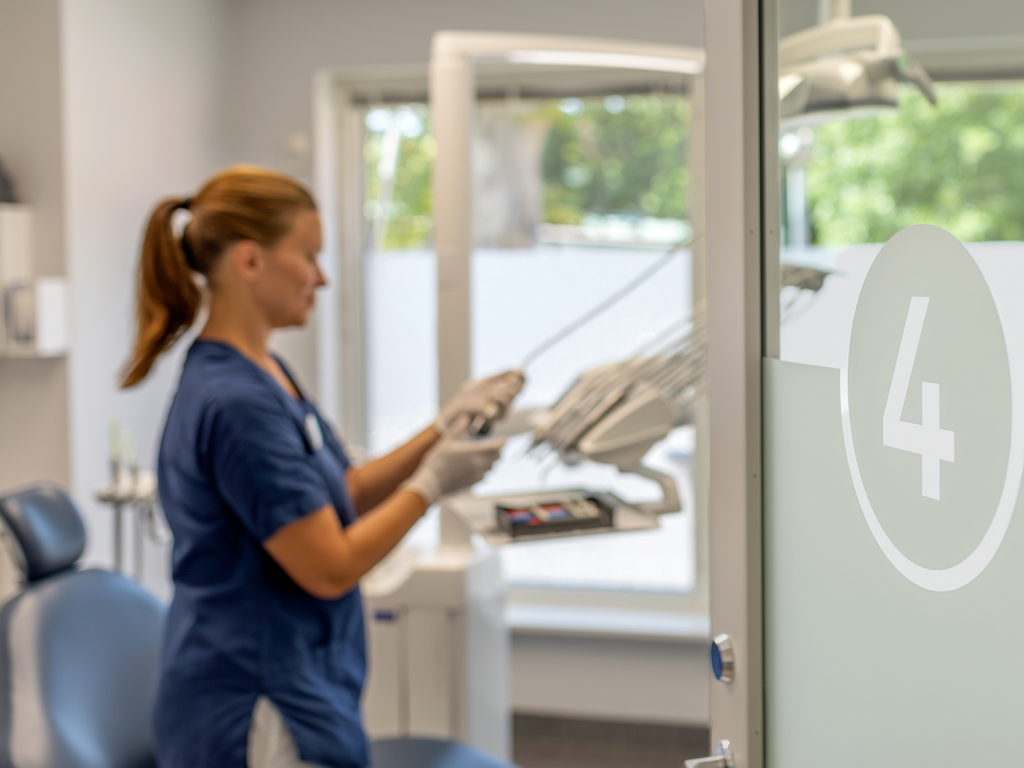 En tandvårdsklinik på Smile Tandvård i Lund där en tandhygienist i blå arbetskläder förbereder en tandvårdsstol och utrustning i ett behandlingsrum märkt med siffran 4. Rummet har stora fönster som släpper in naturligt ljus och ger en ljus och luftig atmosfär.