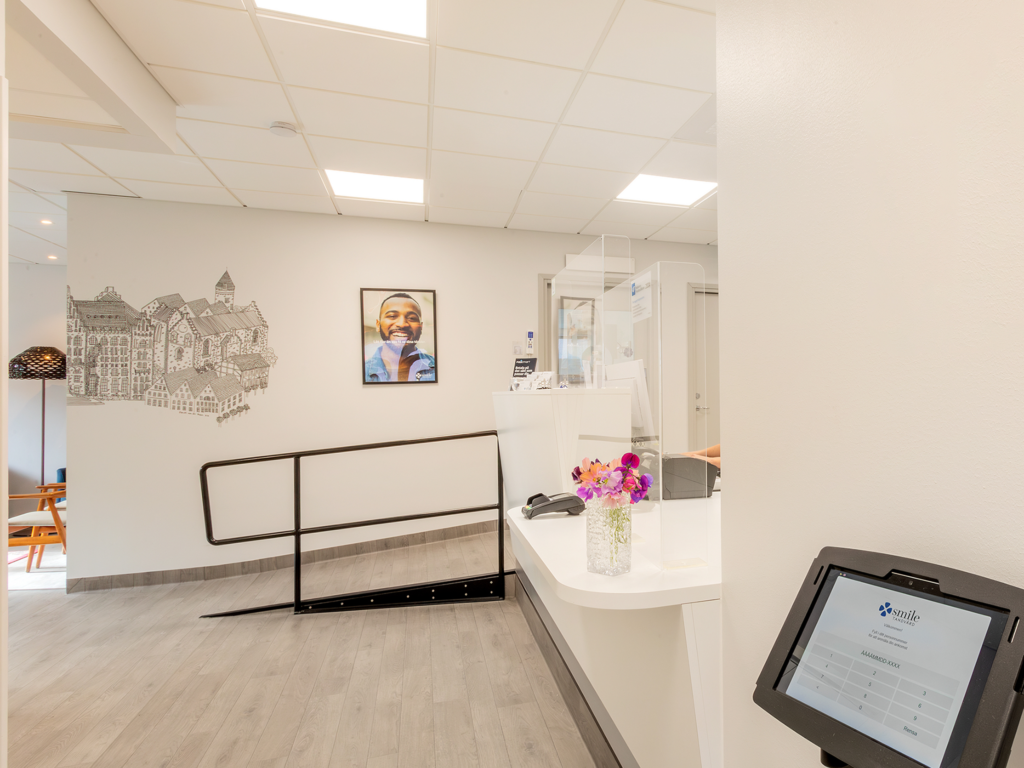 Interiör av receptionen på Smile Tandvård i Lund. Rummet har vita väggar och tak med inbyggd belysning. Till höger syns en digital incheckningsskärm, och längre in finns receptionsdisken med blomsterarrangemang. Väggarna är dekorerade med konstverk och en textlogotyp.