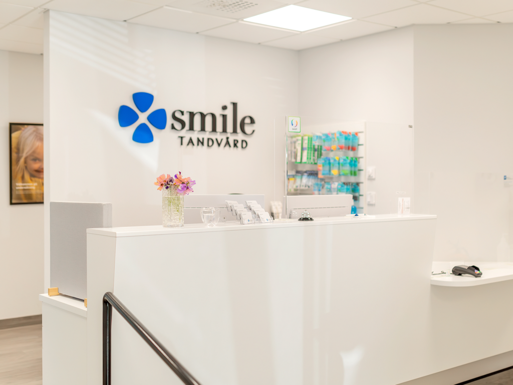 Receptionen på Smile Tandvård i Lund. Disken är vit och bakom den syns klinikens logotyp på väggen, bestående av en blå blomliknande symbol och texten 'Smile Tandvård'. På disken står en vas med blommor och en dator. Väggarna är dekorerade med konstverk och en textlogotyp.