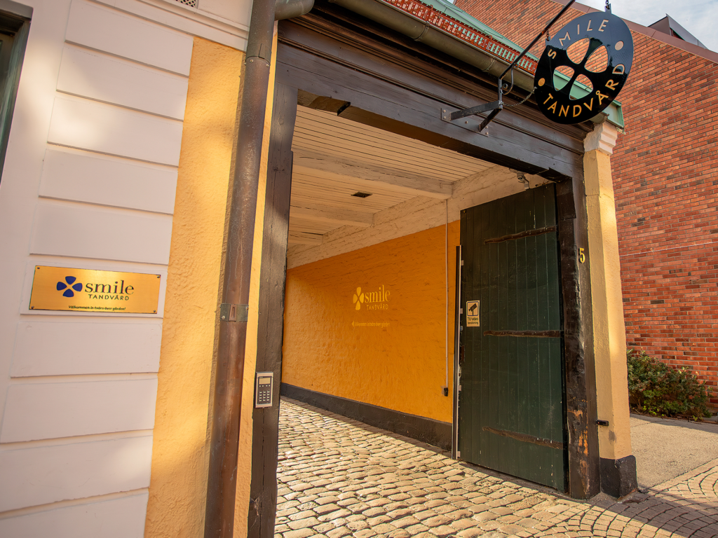 Ingången till Smile Tandvård i Lund, med en öppen port som leder till en gulmålad korridor. På byggnadens utsida syns en svart och vit tandvårdsskylt. En gul pilskylt pekar in mot ingången. Röd tegelarkitektur syns i bakgrunden, under en blå himmel.
