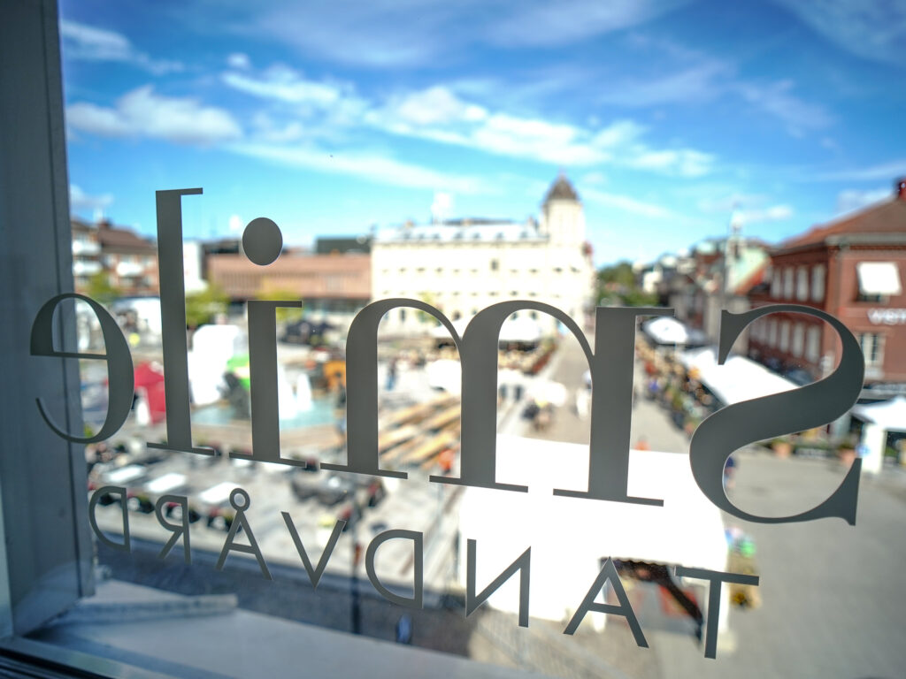 Vy från Smile Tandvård genom ett fönster med logotyp, med utsikt över ett torg och stadsmiljö på en solig dag.