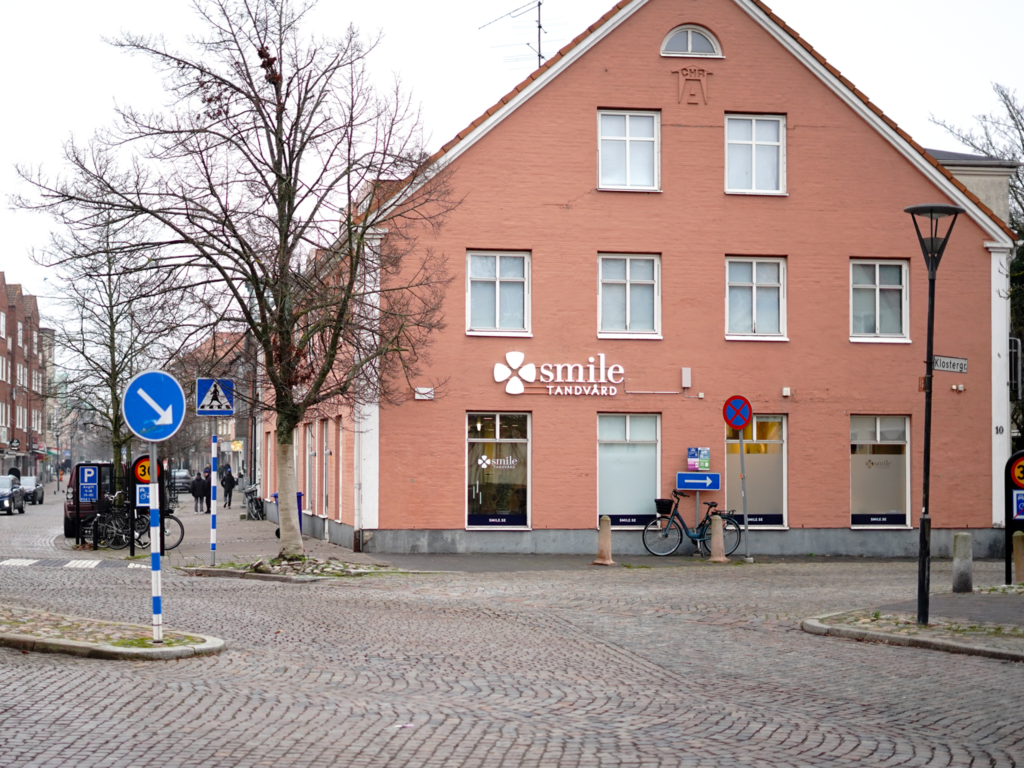 Fasad av Smile Tandvård Trelleborg – Tegelröd byggnad med stora fönster och klinikens logotyp, belägen vid en kullerstensgata i centrala Trelleborg.