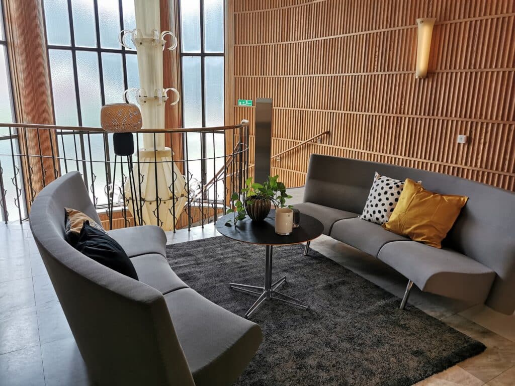 En avslappnande lounge i ett tandläkarväntrum med ett stort panoramafönster som ger utsikt över himlen. Rummet är smakfullt inrett med en stor grå soffa, kompletterad av en rund svart bord och en dekorativ kudde. Bakgrunden visar en modern träpanelvägg och elegant belysning, vilket ger en varm och välkomnande atmosfär.