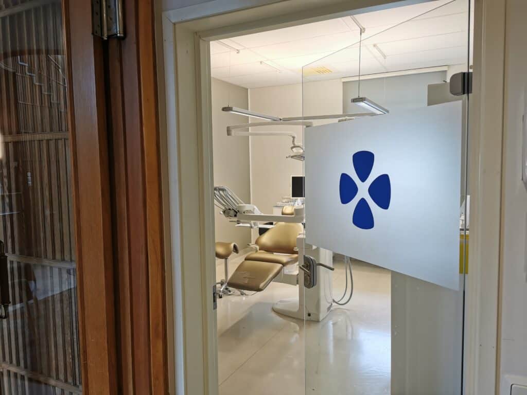 En titt genom en halvöppen dörr in i ett tandvårdsbehandlingsrum med modern utrustning. En tandläkarstol är placerad i mitten av rummet, omgiven av kliniskt vita väggar. Dörren pryds av Smile Tandvårds karakteristiska blå logo, vilket indikerar klinikens varumärkesidentitet.