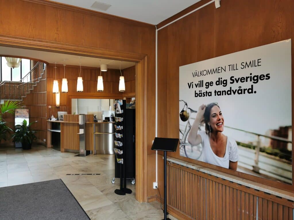Receptionsområdet på Smile Tandvård, med en stor välkomnande skylt som lyder 'Välkommen till Smile, vi vill ge dig Sveriges bästa tandvård.' En mörk receptionsdisk står framför en träpanelvägg, och ett desinfektionsställ för händer är placerat i förgrunden, vilket framhäver klinikens engagemang för kundernas välbefinnande och hygien..