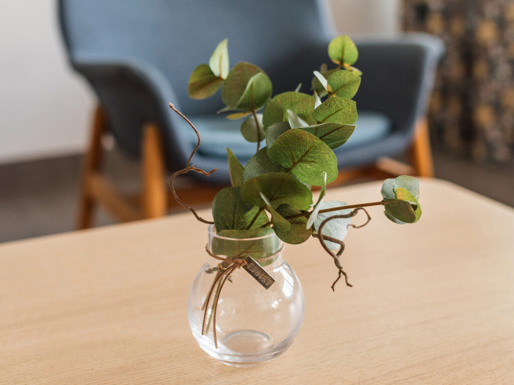Grön kvist i liten glasvas på bord i modern lounge med blå fåtölj i bakgrunden.