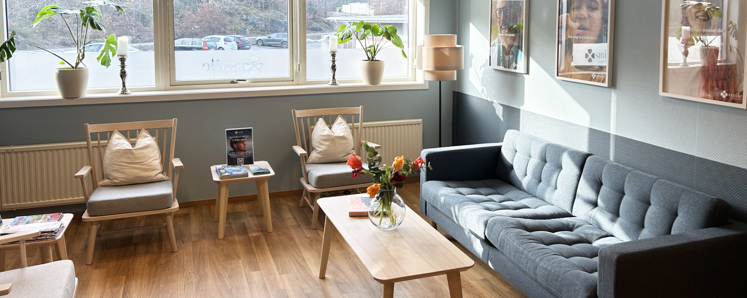 Bilden föreställer interiören hos Smile Tandvård i Torslanda, en modern och inbjudande tandläkarmottagning. Utrymmet är designat för att ge en avkopplande atmosfär till patienter med sin ljusa och öppna planlösning. I bakgrunden syns receptionsdisk där patienter välkomnas. Inredningen inkluderar en elegant ljuskrona, en svart soffa och ett kaffebord, vilket ger en hemtrevlig känsla. Hyllorna och konstverken på väggarna bidrar till en lugnande miljö.