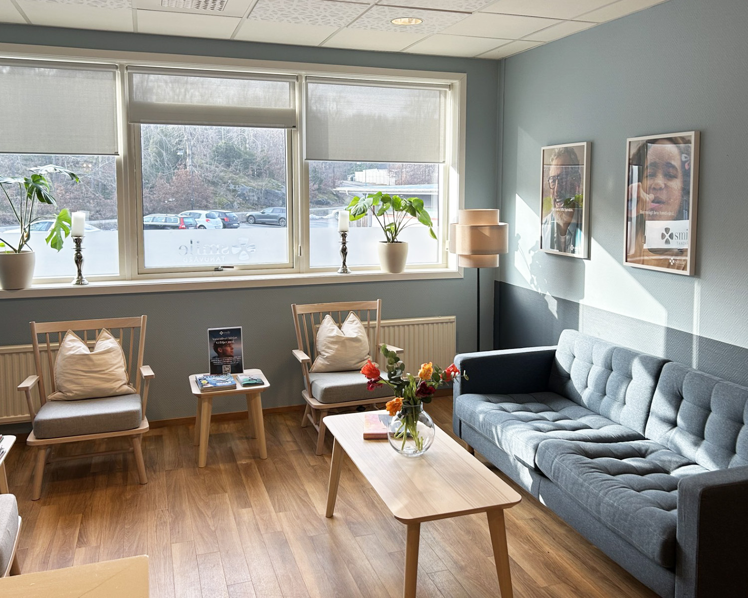 Bilden föreställer interiören hos Smile Tandvård i Torslanda, en modern och inbjudande tandläkarmottagning. Utrymmet är designat för att ge en avkopplande atmosfär till patienter med sin ljusa och öppna planlösning. I bakgrunden syns receptionsdisk där patienter välkomnas. Inredningen inkluderar en elegant ljuskrona, en svart soffa och ett kaffebord, vilket ger en hemtrevlig känsla. Hyllorna och konstverken på väggarna bidrar till en lugnande miljö.