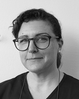 Svartvitt porträtt av en leende person med mörkt hår uppsatt bakåt och glasögon med mörk båge. Personen bär en mörk tröja och tittar mot kameran mot en ljus, neutral bakgrund.