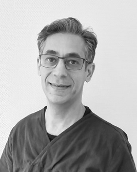 Porträtt av en manlig tandhygienist med glasögon och mörkt kort hår, leende mot kameran, iförd en mörk arbetsdräkt med en namnbricka, mot en neutral bakgrund.