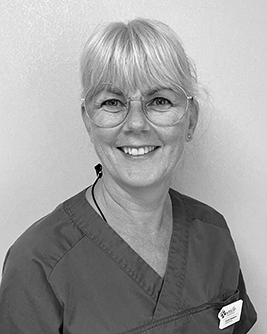 Porträtt av en leende kvinnlig tandsköterska med glasögon och ljust uppsatt hår. Hon bär mörka tandvårdskläder och ser direkt in i kameran framför en enkel vit bakgrund vilket ger ett professionellt och tillmötesgående intryck.