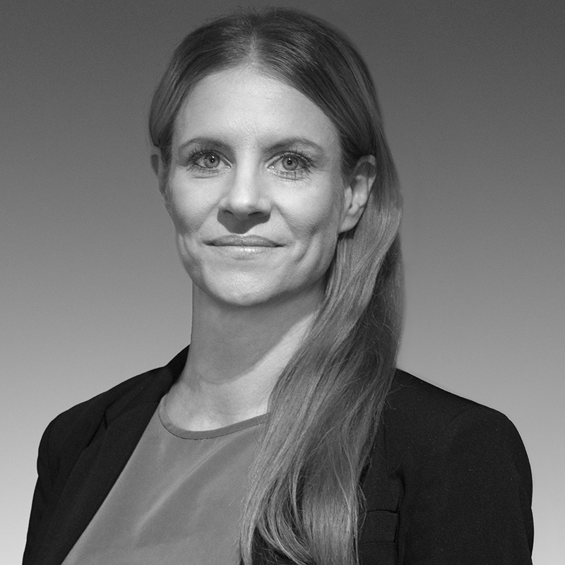 Svartvit bild av en teamservice medlem med långt, rakt hår, neutralt ansiktsuttryck, iklädd en mörk blazer över en ljus topp.