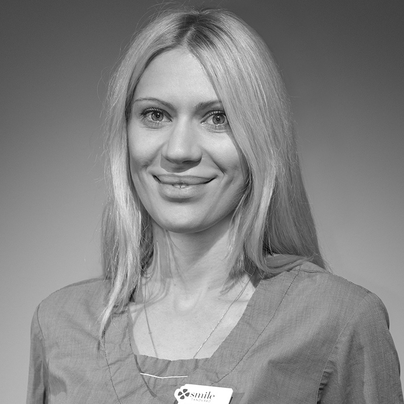 Svartvit bild av en kvinnlig tandhygienist med långt blont hår och ett vänligt leende, bär en namnskylt och en tandhygienistuniform.