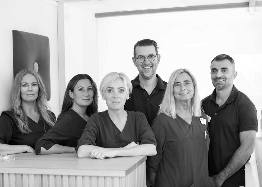 Gruppbild av leende och professionella tandläkare, tandsköterskor och tandhygienister på Smile Tandvård Kristianstad tandläkarklinik, klädda i sina blå arbetskläder och stående i klinikens receptionsområde.