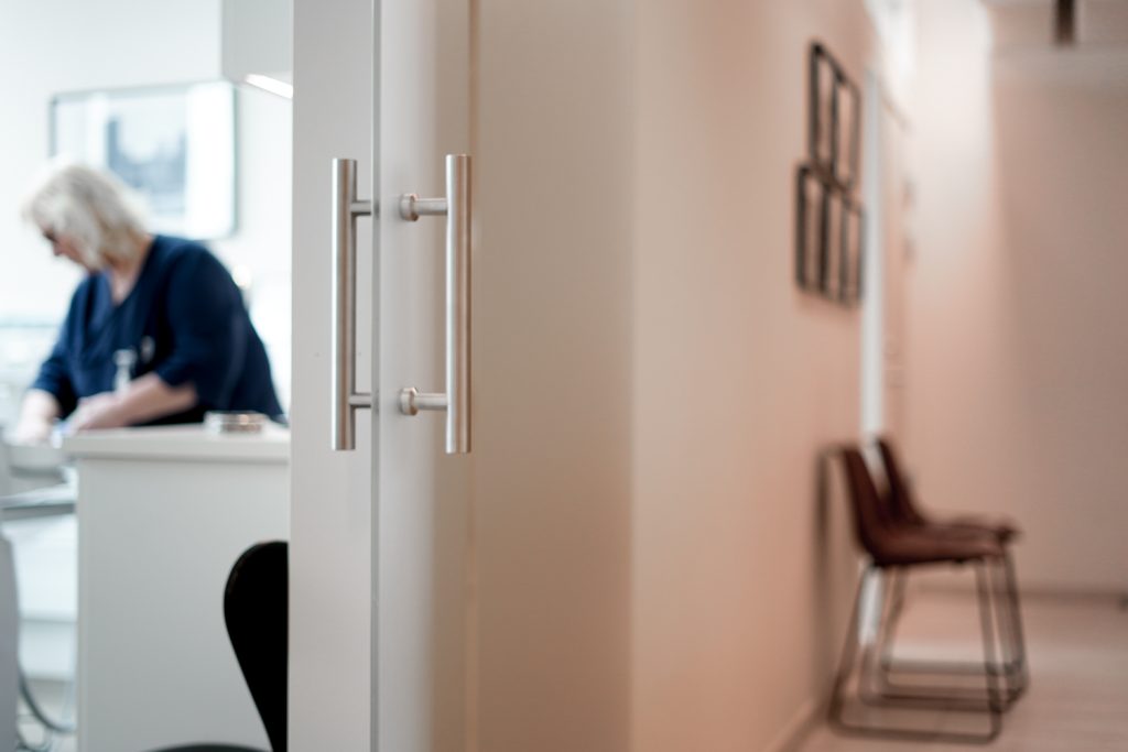 Bildens fokus ligger på en stol av läder vid ingången till ett behandlingsrum hos Smile Tandvård Göteborg Avenyn, med suddig bakgrund där en tandvårdspersonal arbetar vid receptionen.