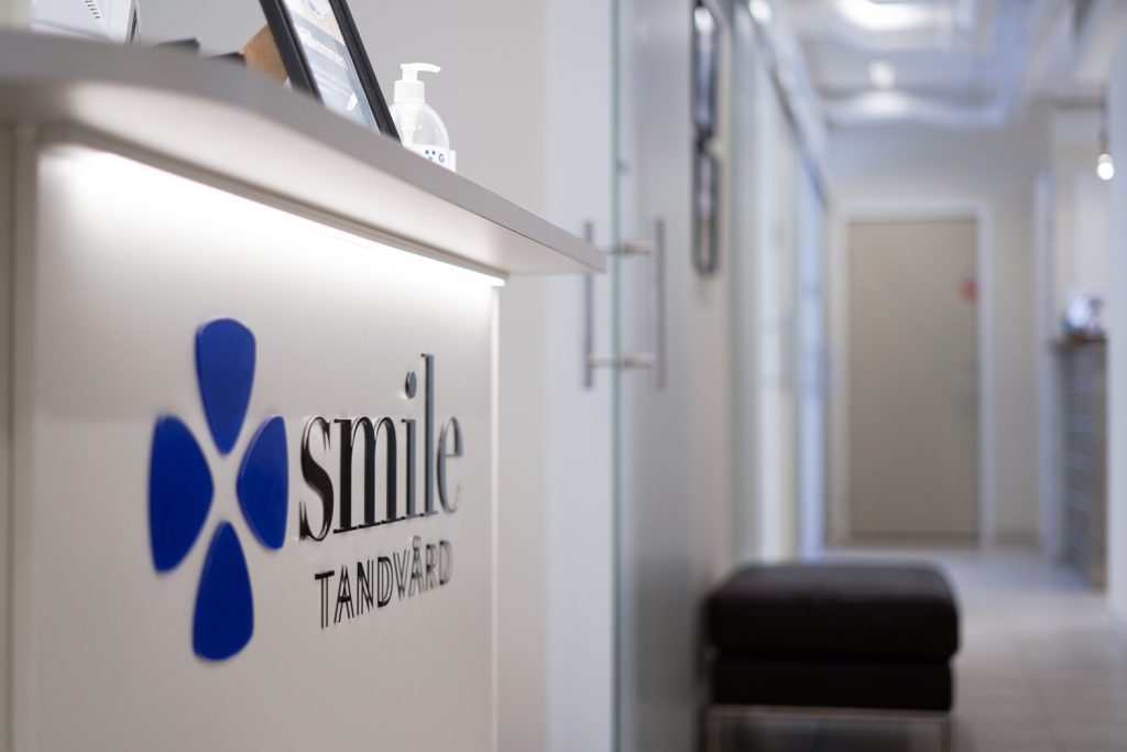 En korridor på Smile Tandvård med företagets logotyp i förgrunden på en upplyst receptionsdisk och en handdesinfektionsdispenser ovanpå, med en inbjudande passage som leder till behandlingsrummen i bakgrunden.