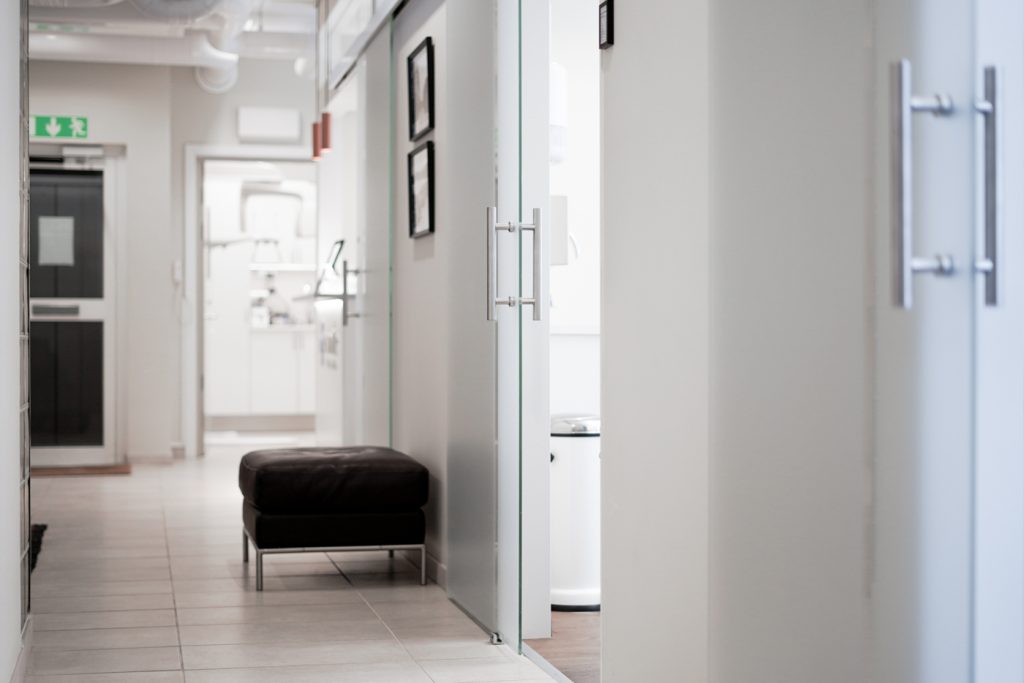 Ljus och modern korridor på Smile Tandvård Avenyn med behandlingsrum åt båda sidor, utrustade med transparenta dörrar och diskreta skyltar, samt en bekväm sittbänk placerad längs väggen.