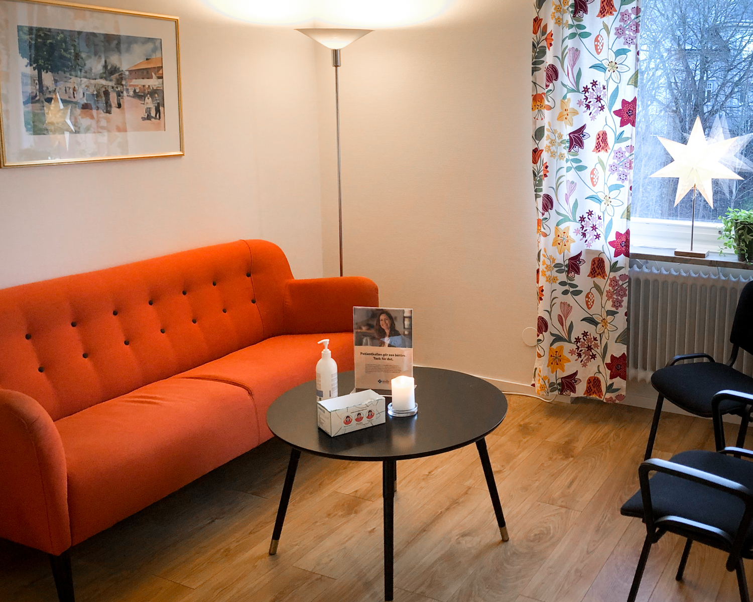 En välkomnande väntrumsmiljö hos Smile Tandvård i Molkom med en orange soffa, ett runt kaffebord med en informationsbroschyr och två handdesinfektionsflaskor, mot en vägg med ett livfullt blommönster och en intillstående svart stol.