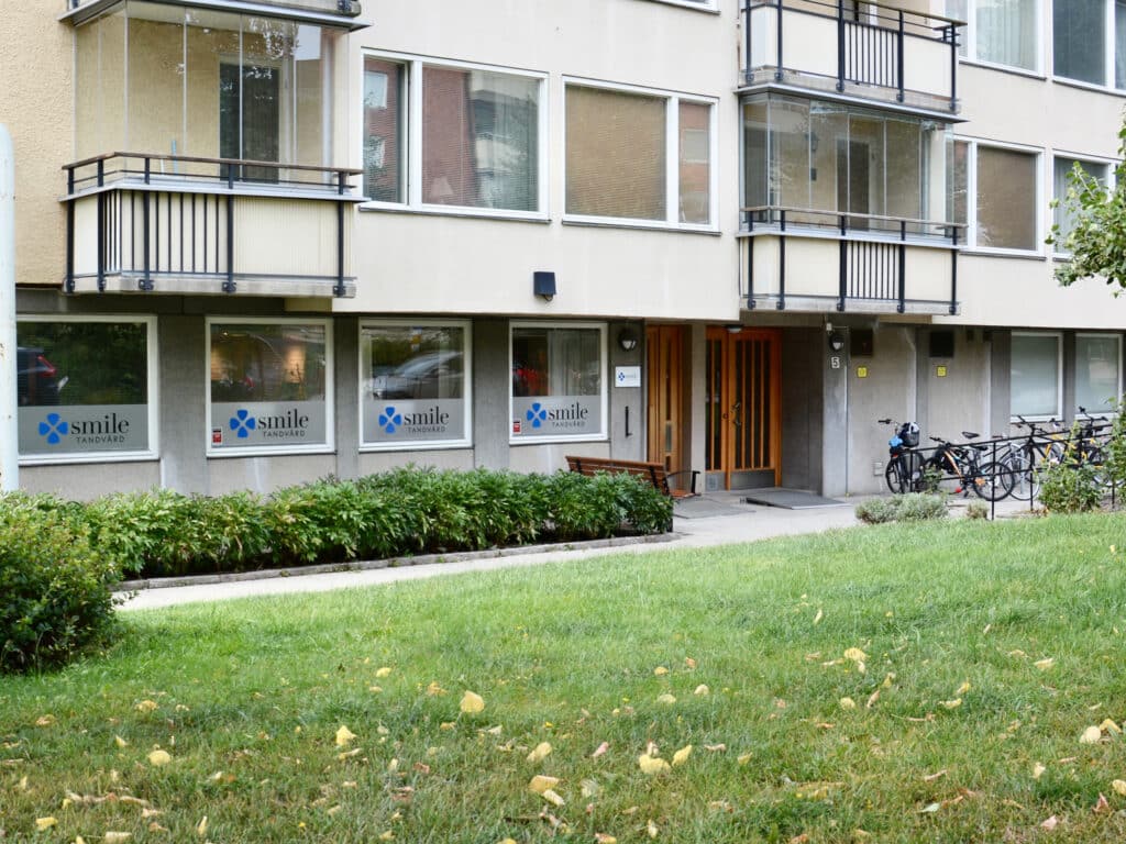 Exteriör av Smile Tandvård i Solna, med klinikens fönster och entré i ett flerfamiljshus, omgiven av grönska och cykelställ.