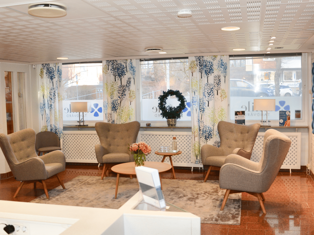 Väntrum på Smile Tandvård Solna – en ljus och inbjudande miljö med bekväma fåtöljer, ett runt soffbord med en vas fylld med rosa blommor och stora fönster som släpper in naturligt ljus. Dekorativa gardiner och en grön krans på fönstret skapar en varm och välkomnande atmosfär