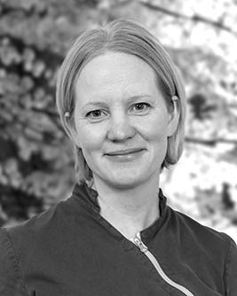 Porträtt av en kvinnlig tandsköterska med ett mjukt leende och kort hår, iförd en mörkfärgad tandvårdsuniform med dragkedja, mot en bakgrund av oskarp grönska.