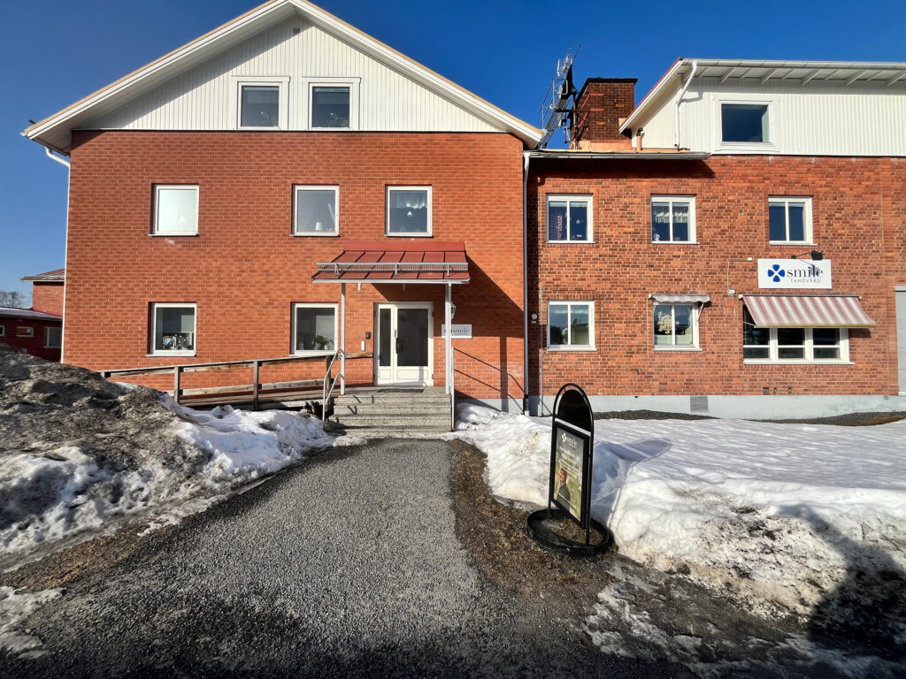 Entrén till Smile Tandvård Höga Kusten i en röd tegelbyggnad med gångväg fram till kliniken och snö på marken.