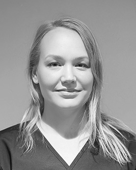 Porträtt i svartvitt av en kvinnlig tandsköterska med ljust långt hår, klädd i mörka arbetskläder, som tittar rakt in i kameran med ett vänligt ansiktsuttryck.
