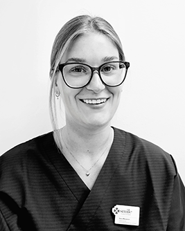 Svartvitt porträtt av en leende person med ljust hår uppsatt bakåt och glasögon med mörk båge. Personen bär en mörk tröja och tittar mot kameran mot en ljus, neutral bakgrund.