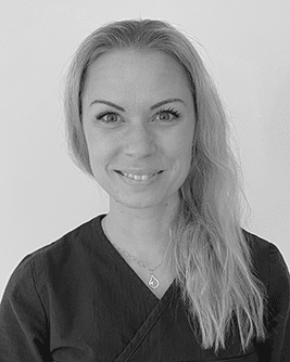 Porträtt av en leende kvinna med långt blont hår, klädd i mörka arbetskläder, stående mot en ljus bakgrund.