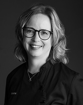 Porträtt av tandsköterska, kvinna med glasögon och lockigt hår, leende, svart arbetsklädsel.