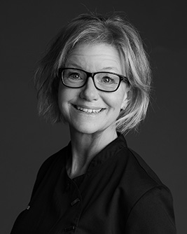 Porträtt av tandsköterska, kvinna med glasögon, leende, svart arbetsklädsel.