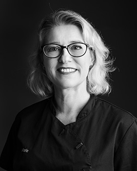 Porträtt av tandsköterska, kvinna med glasögon, kort hår, leende, svart arbetsklädsel.