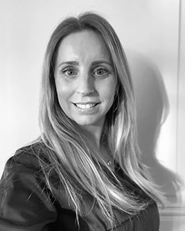 Svartvitt porträtt av en tandhygienist med långt ljust hår. Personen bär en mörk tröja och tittar mot kameran mot en ljus, neutral bakgrund.