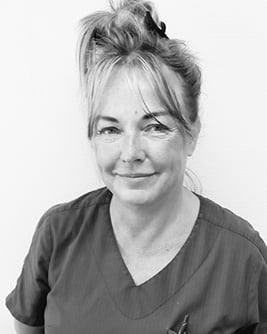 Svartvitt porträtt av en leende tandsköterska med ljust kort hår. Personen bär en mörk tröja och tittar mot kameran mot en ljus, neutral bakgrund.