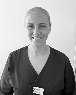 Svartvitt porträtt av en leende tandhygienist med ljust uppsatt hår. Personen bär en mörk tröja och tittar mot kameran mot en ljus, neutral bakgrund.