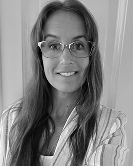 Svartvitt porträtt av en leende person med ljust mörkt hår och glasögon med ljus båge. Personen bär en mörk tröja och tittar mot kameran mot en ljus, neutral bakgrund.