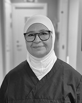 Svartvitt porträtt av en leende person med långt mörkt hår och glasögon med ljus båge. Personen bär en mörk tröja och tittar mot kameran mot en ljus, neutral bakgrund.