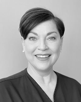 Svartvitt porträtt av en leende tandhygienist med kort mörkt hår. Personen bär en mörk tröja och tittar mot kameran mot en ljus, neutral bakgrund.