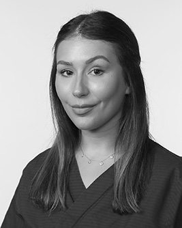 Svartvitt porträtt av en leende ortodontiassistent med långt mörkt hår. Personen bär en mörk tröja och tittar mot kameran mot en ljus, neutral bakgrund.