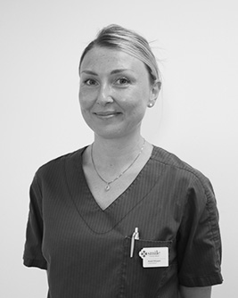 Svartvitt porträtt av en leende tandhygienist med ljust uppsatt hår. Personen bär en mörk tröja och tittar mot kameran mot en ljus, neutral bakgrund.