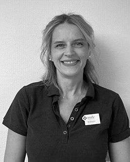 Svartvitt porträtt av en leende tandhygienist med ljust uppsatt hår. Personen bär en mörk tröja och tittar mot kameran mot en ljus, neutral bakgrund.