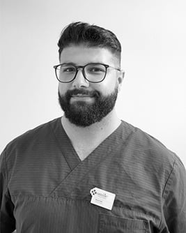 Svartvitt porträtt av en leende manlig tandhygienist med kort mörkt hår, glasögon och skägg.