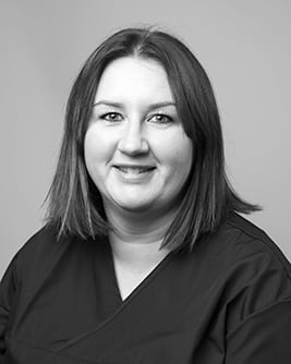 Svartvitt porträtt av en leende tandsköterska med mörkt medellångt hår. Personen bär en mörk tröja och tittar mot kameran mot en ljus, neutral bakgrund.