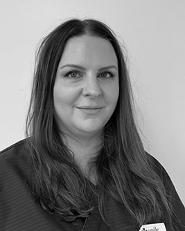 Svartvitt porträtt av en tandläkare med långt mörkt hår. Personen bär en mörk tröja och tittar mot kameran mot en ljus, neutral bakgrund.