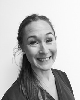 Svartvitt porträtt av en leende tandhygienist med ljust uppsatt hår. Personen bär en mörk tröja och tittar mot kameran mot en ljus, neutral bakgrund.